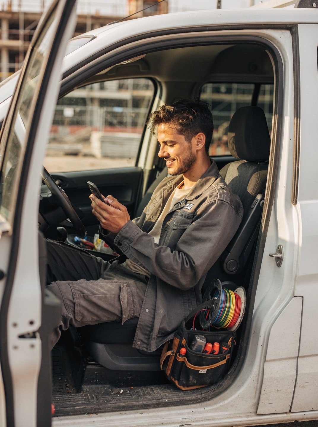 A young electrician smiles at his phone in the driver’s seat of a work van